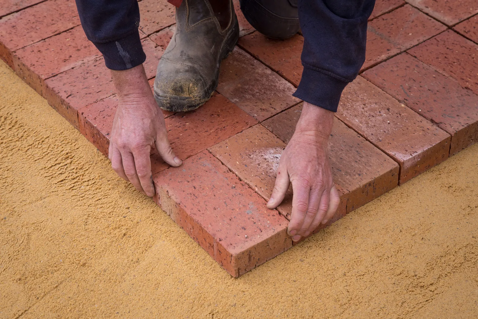 Hands laying paver blocks into a sand bed in a herringbone pattern